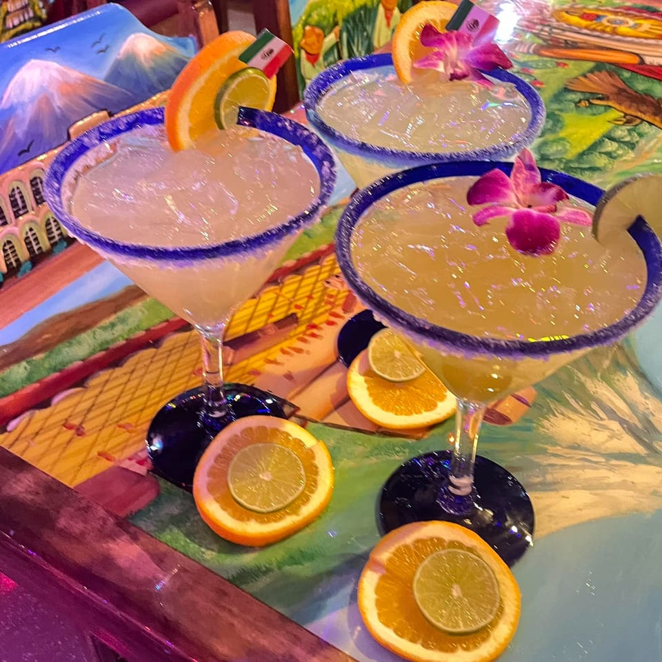 Three margaritas with orange and lime slices, some with orchids, on a colorful Mexican-themed table.