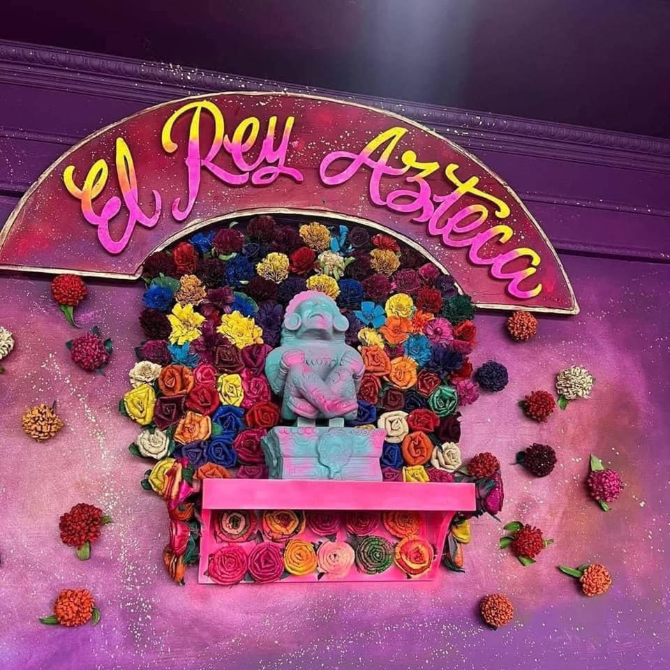 El Rey Azteca sign with a blue and pink Aztec statue surrounded by colorful fabric flowers.