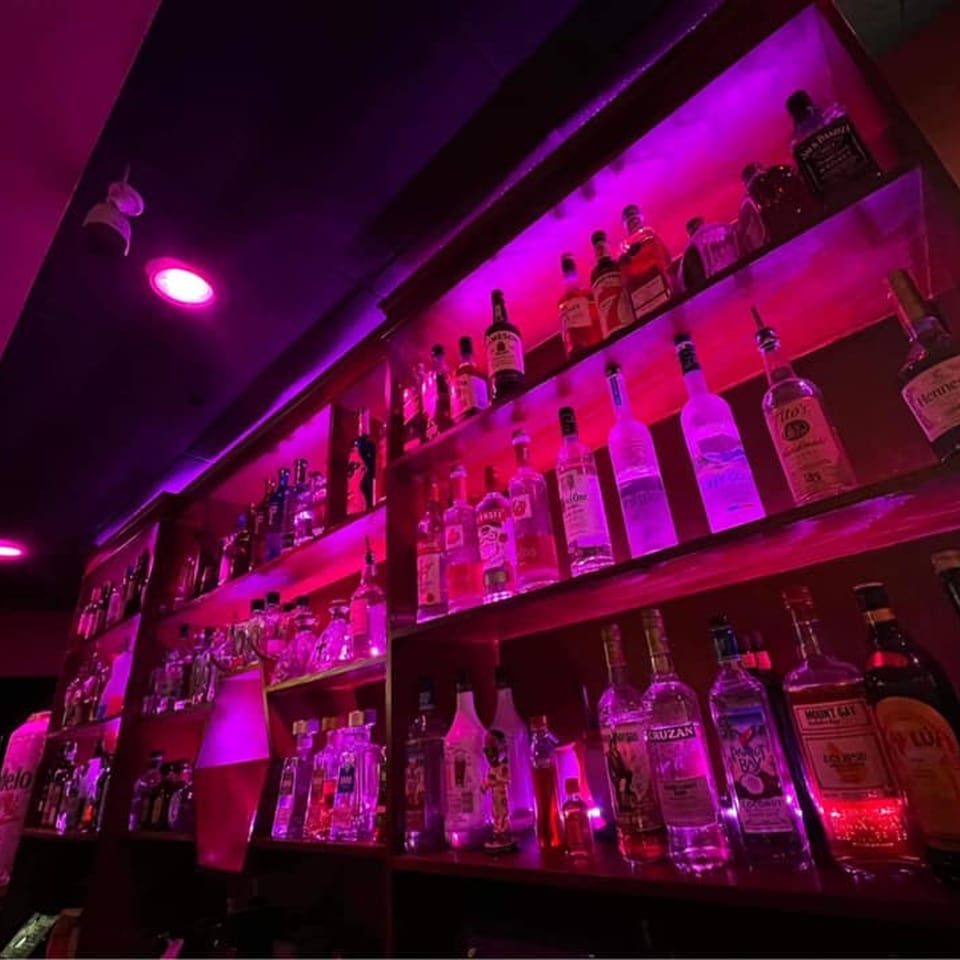 A dimly lit bar shelf at El Rey Azteca, showcasing various liquor bottles under vibrant pink lighting.
