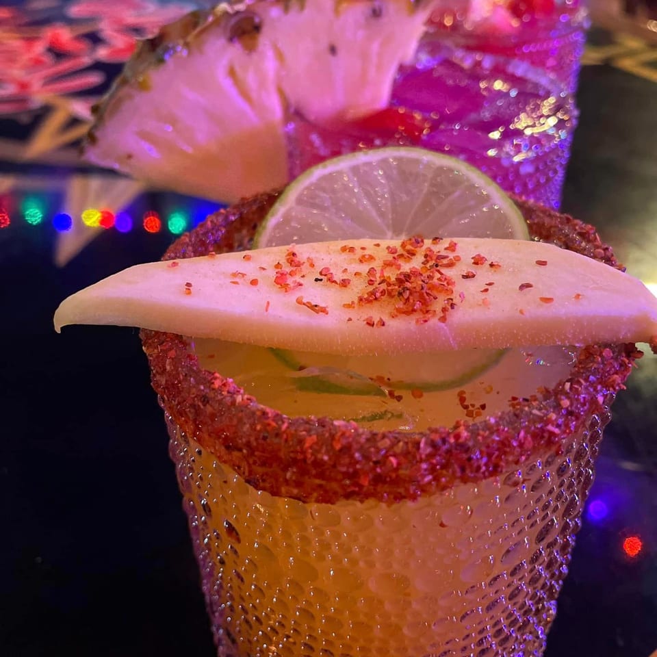 A Mexican cocktail with a chili-lime rim, garnished with a lime slice and a mango slice.